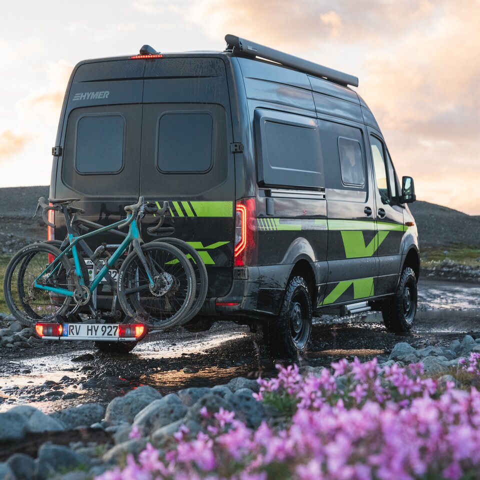 Sprinter-4x4-bobilen HYMER Grand Canyon S CrossOver står på en ujevn vei med blomster og gress, på sykkelstativet står sykler.