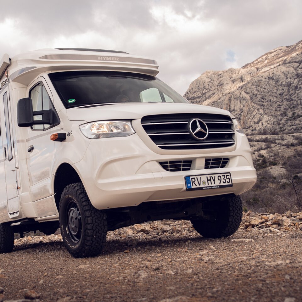 Den hvite Mercedes Allrad-bobilen HYMER ML-T står på en stigende steinvei med klippeåser og lett skyet himmel i bakgrunnen.