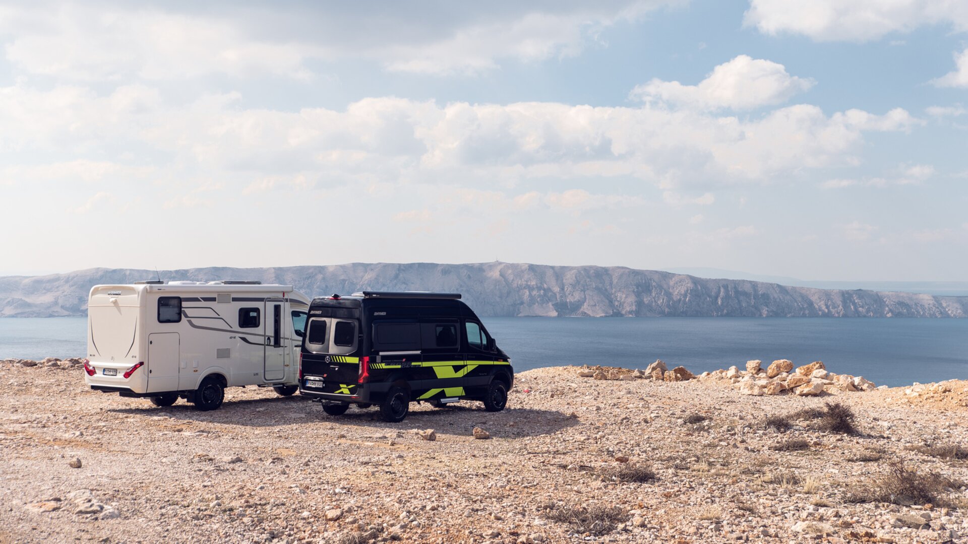 To offroad-bobiler i hvitt samt svart-grønt står på en solbelyst slette over havet, i bakgrunnen er klipper synlige.