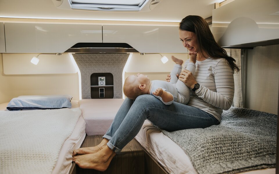 Young woman sits on a bed in the rear of a camper van with a baby on her lap