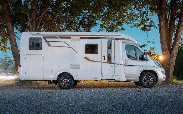 On a gravel area with burning street lamps and a HYMER Exsis-t parked under large trees from the side