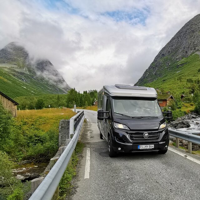 HYMER Exsis-t on a narrow mountain road surrounded by rocky mountains, fir trees, green meadows and a mountain stream
