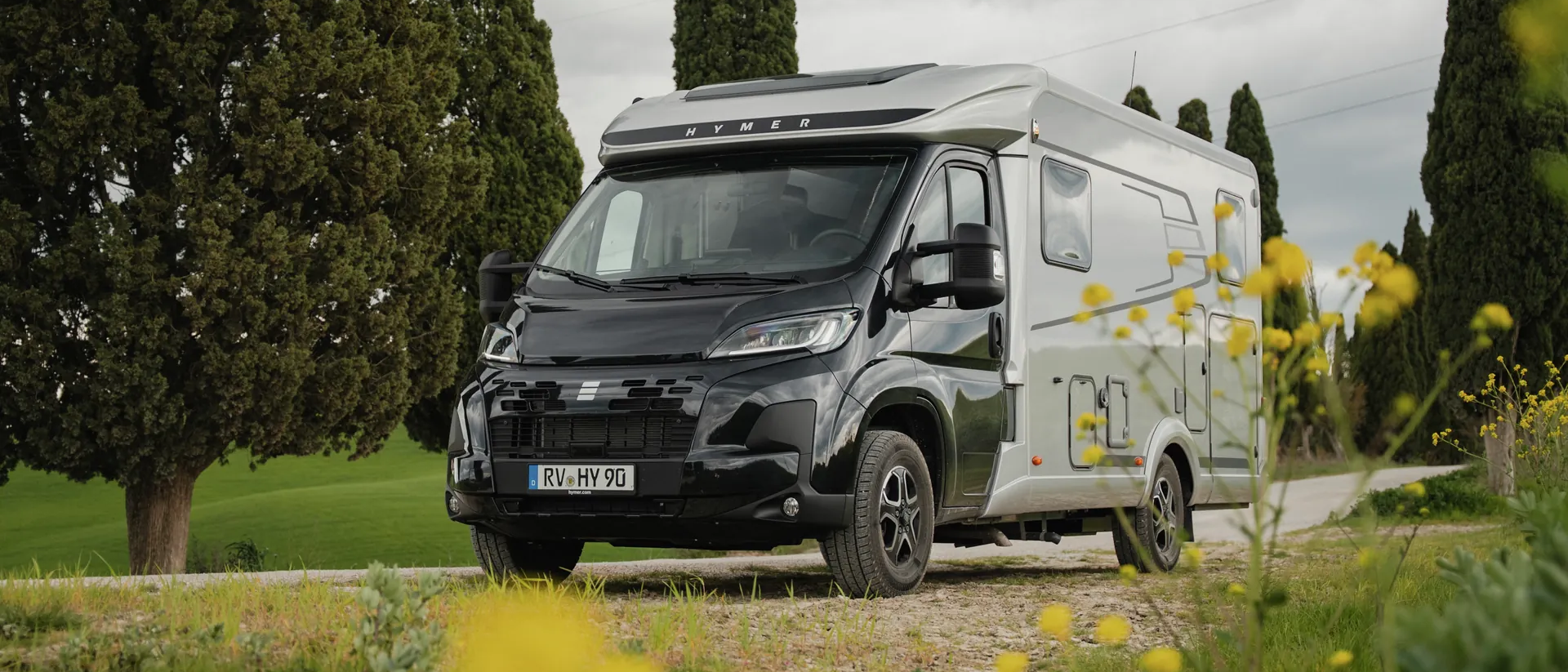 The Hymer Exsis‑t with a black cab and silver body shell is parked on a rural track, surrounded by countryside with cypress trees, flowers in the foreground and a cloudy sky.