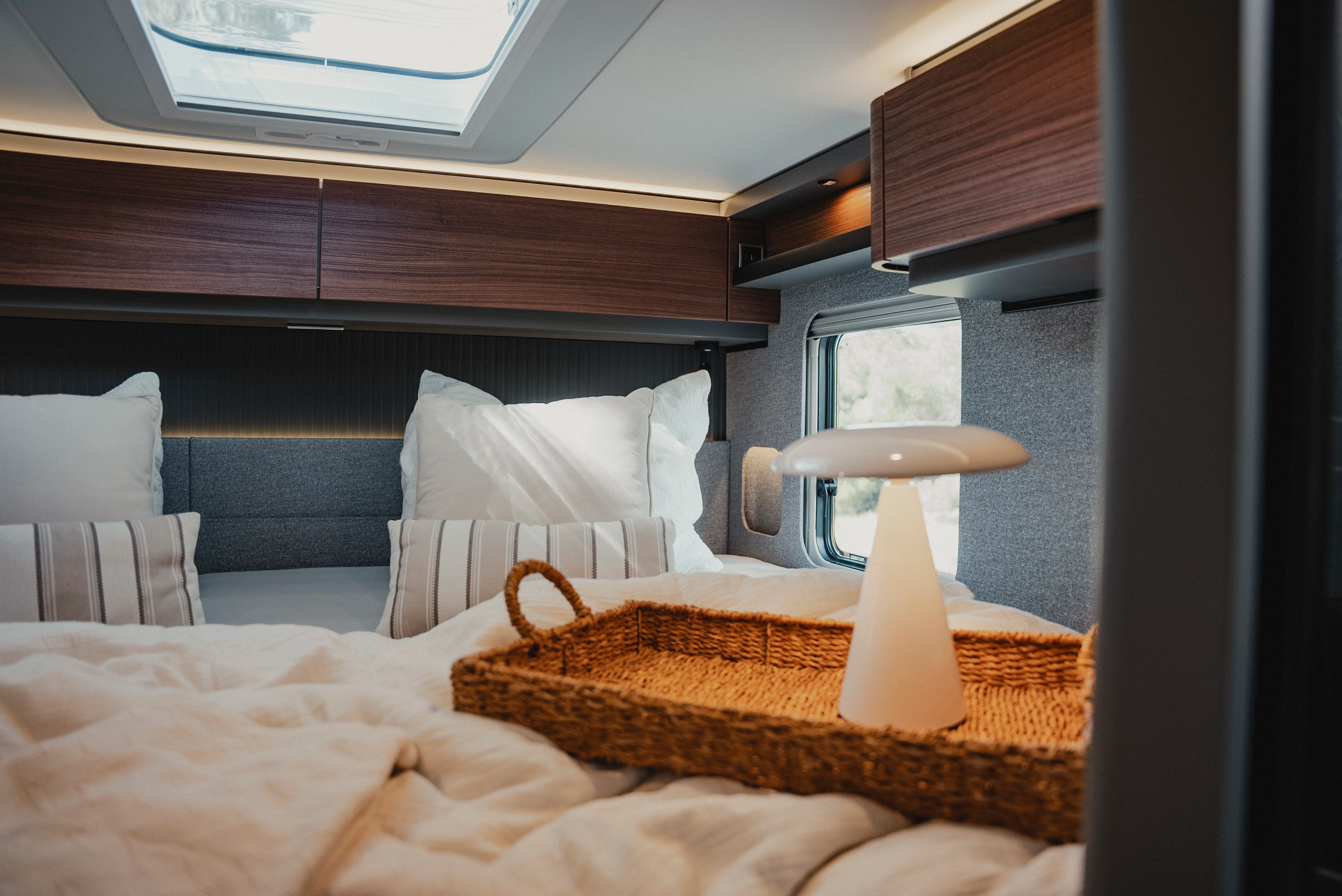 Experience luxurious comfort with a stylishly equipped sleeping area, complemented by modern overhead storage cabinets in wood finish, here in Rochefort walnut. The combination of natural light and warm lighting creates an atmosphere of relaxation and elegance for your travels.