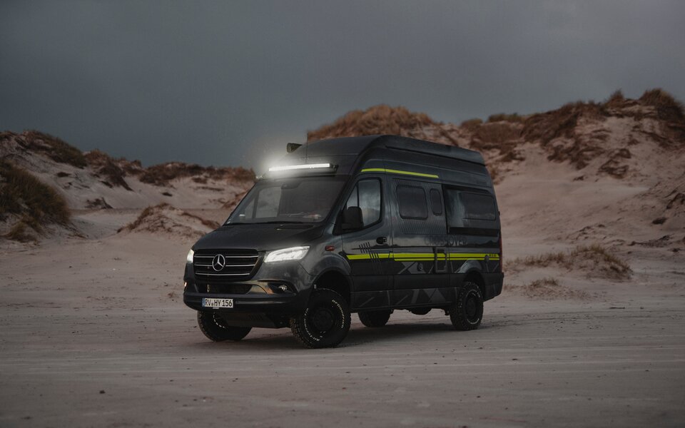 HYMER-Offroad-Camper steht in der Dämmerung mit eingeschaltetem Scheinwerfer und Light-Bar an einem Sandstrand mit Sanddünen im Hintergrund.
